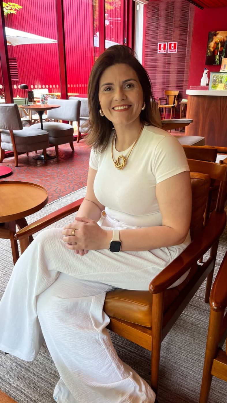 Luana Claudino sentada em um café, de roupa clara, sorrindo